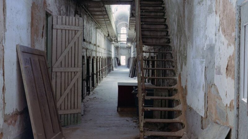 A decaying cellblock corridor inside Eastern State Penitentiary in Philadelphia, with peeling walls, open cell doors, and a vaulted skylight at the far end