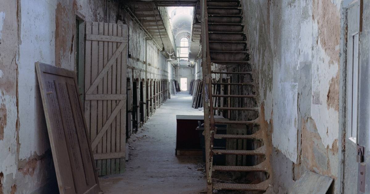 A decaying cellblock corridor inside Eastern State Penitentiary in Philadelphia, with peeling walls, open cell doors, and a vaulted skylight at the far end